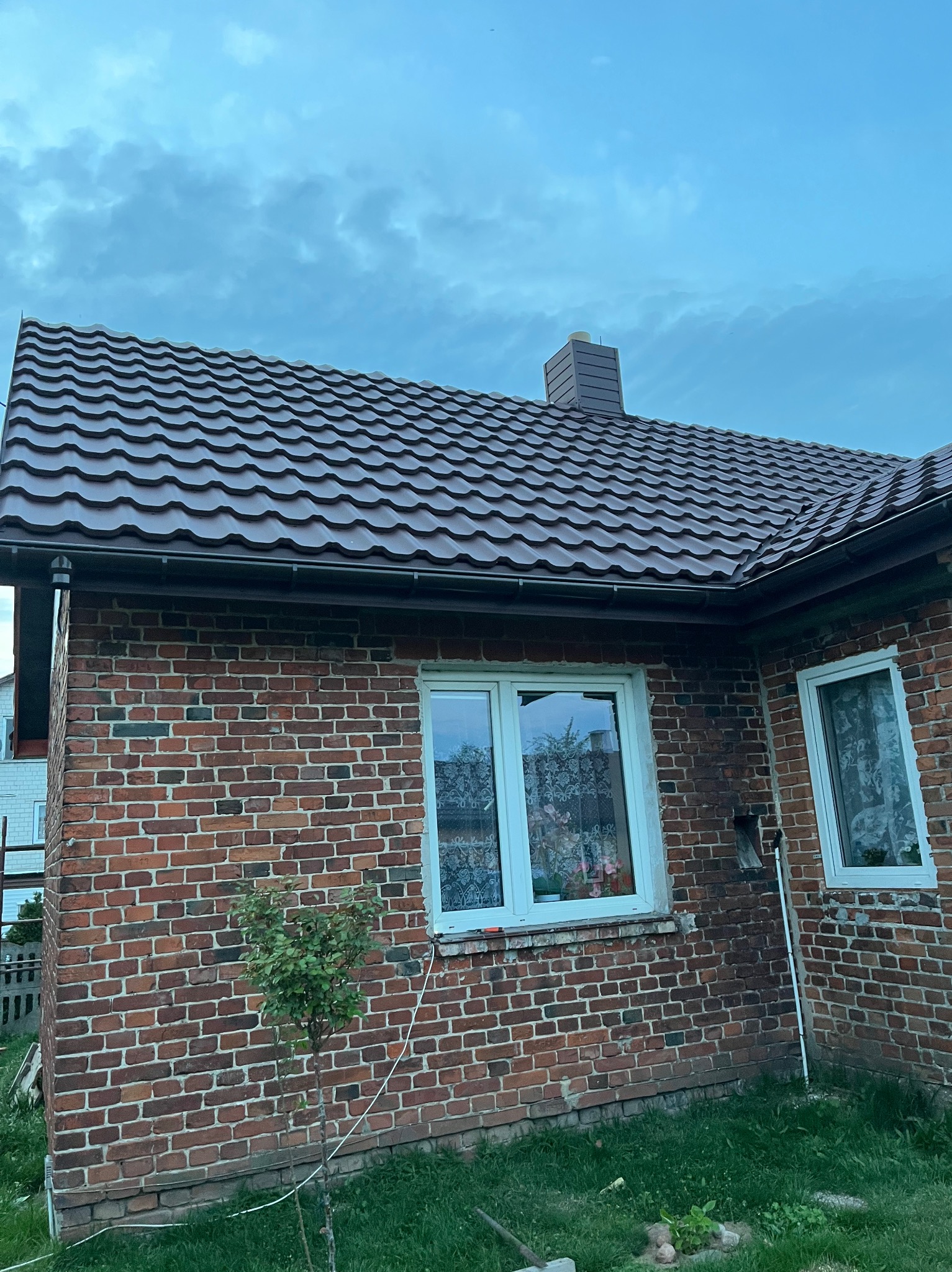 Dom z czerwonej cegły z nowym, brązowym dachem z dachówki ceramicznej. Widoczne rynny w kolorze dachu i komin. Ujęcie z dołu, pochmurne niebo w tle.
