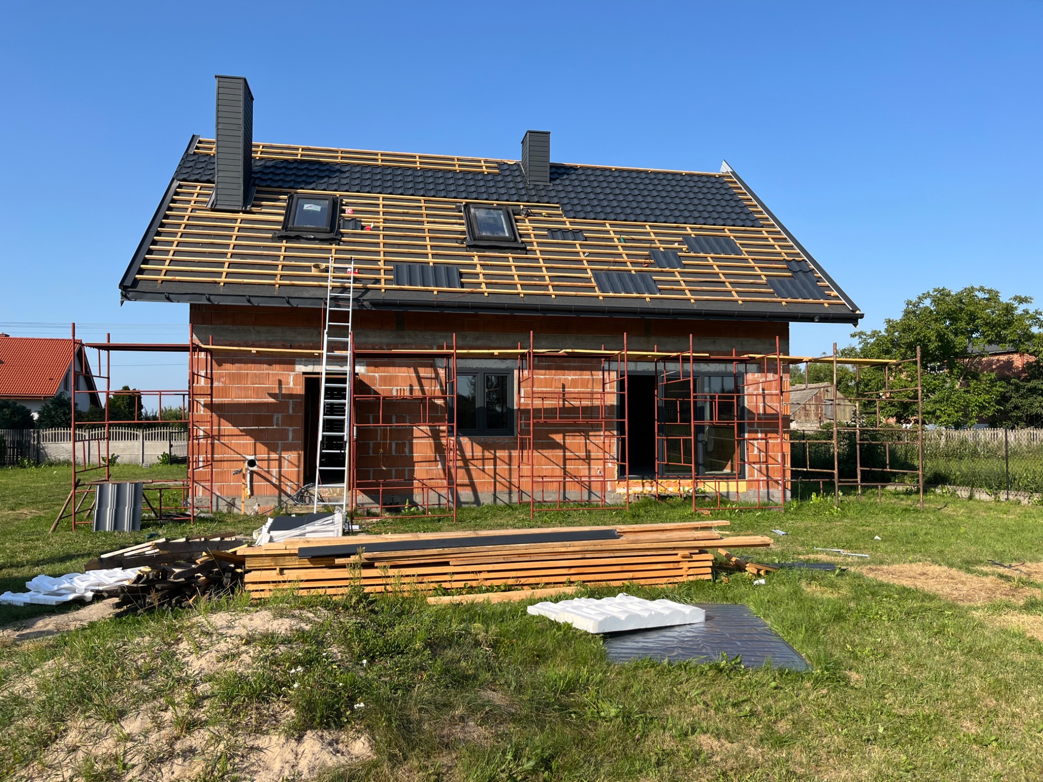 Dom w trakcie budowy z częściowo pokrytym dachem czarną dachówką, widoczne rusztowania, drewno i okna dachowe. Ujęcie w słoneczny dzień.