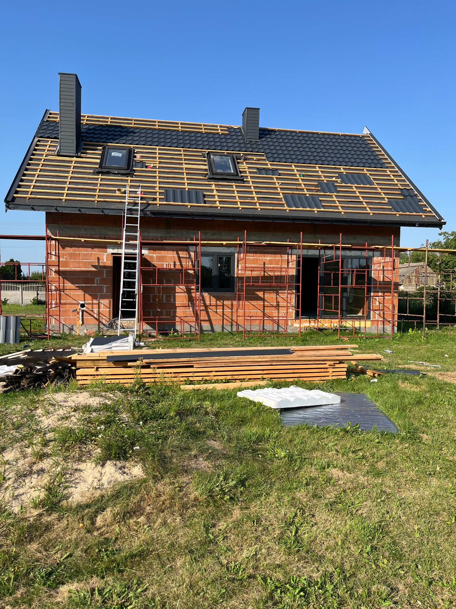 Budowa domu jednorodzinnego w trakcie krycia dachu blachodachówką w kolorze grafitowym. Widoczne rusztowania, drabina i materiały budowlane. Słoneczny dzień.