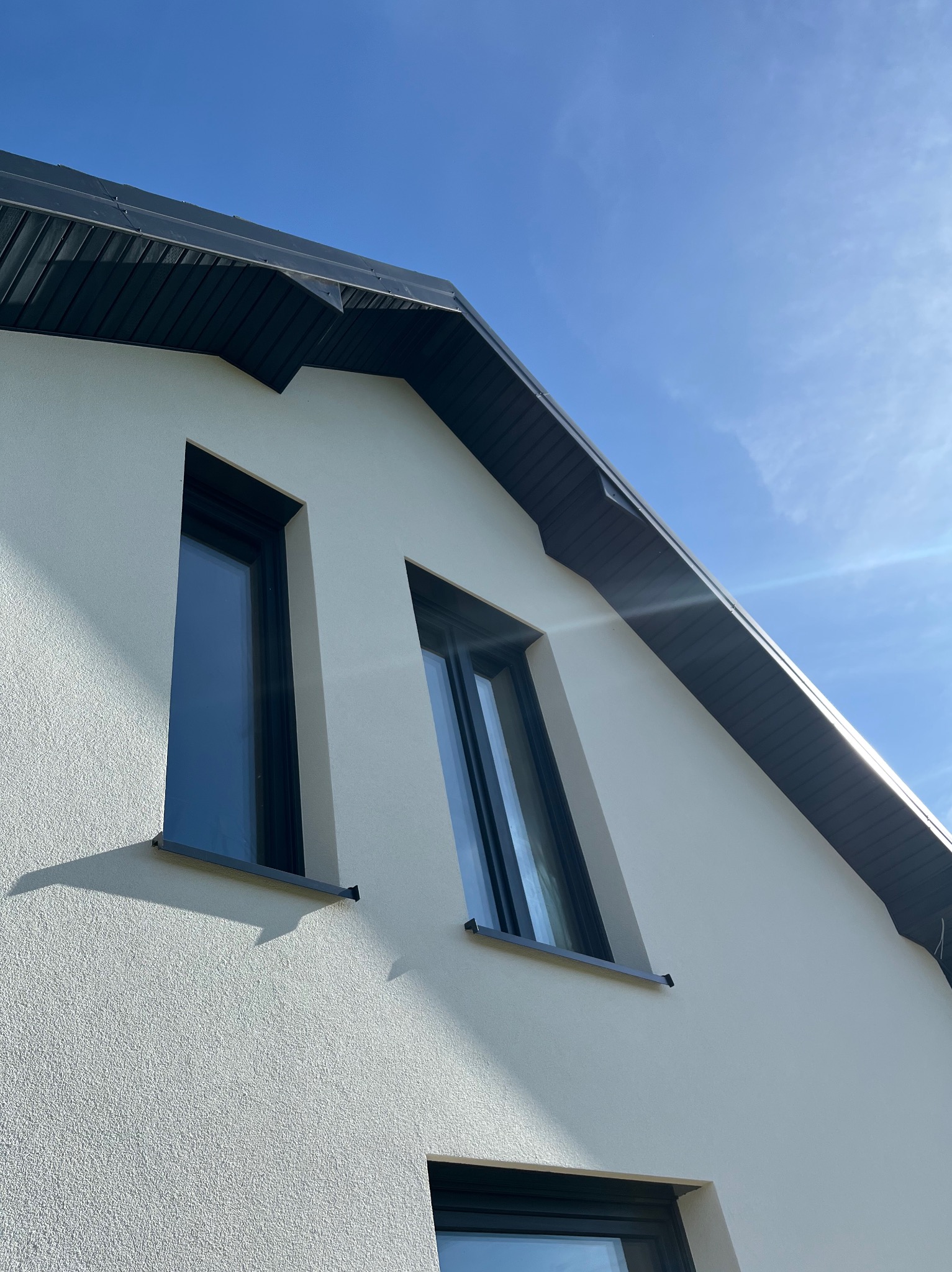 Fragment elewacji domu z oknami i dachem w kolorze antracytu na tle błękitnego nieba. Widok z dołu, podkreślający geometrię budynku i nowoczesny styl.