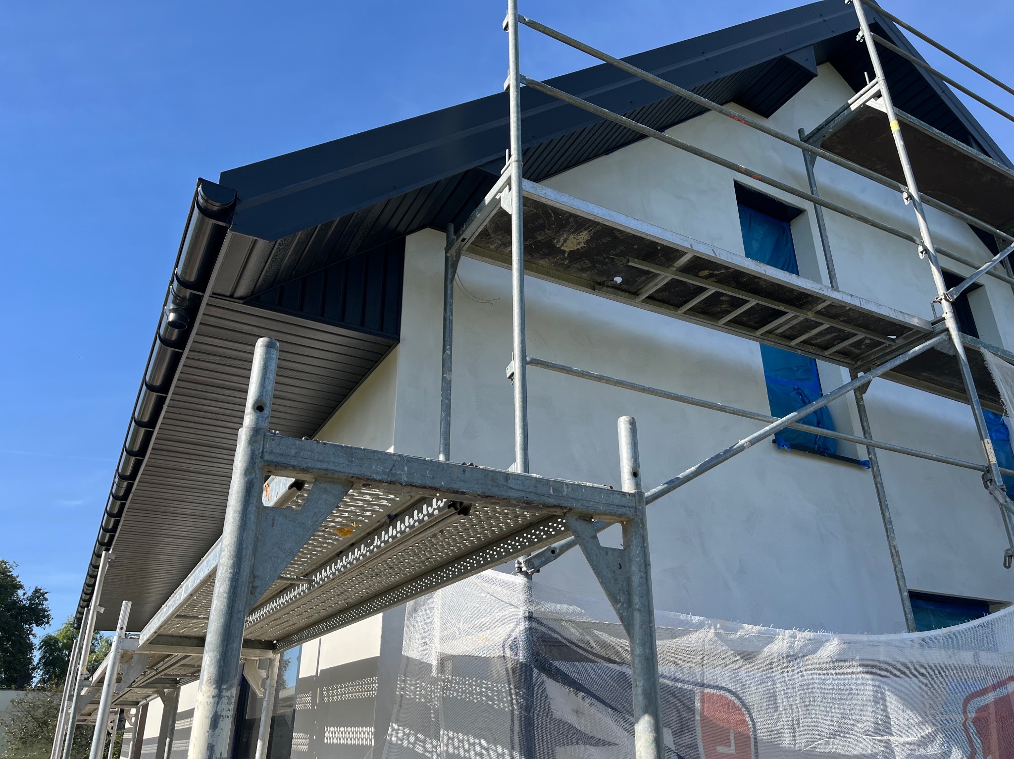 Dom w trakcie prac wykończeniowych: rusztowanie, świeżo otynkowana elewacja, czarne rynny i podbitka dachowa pod błękitnym niebem. Widok z dołu.