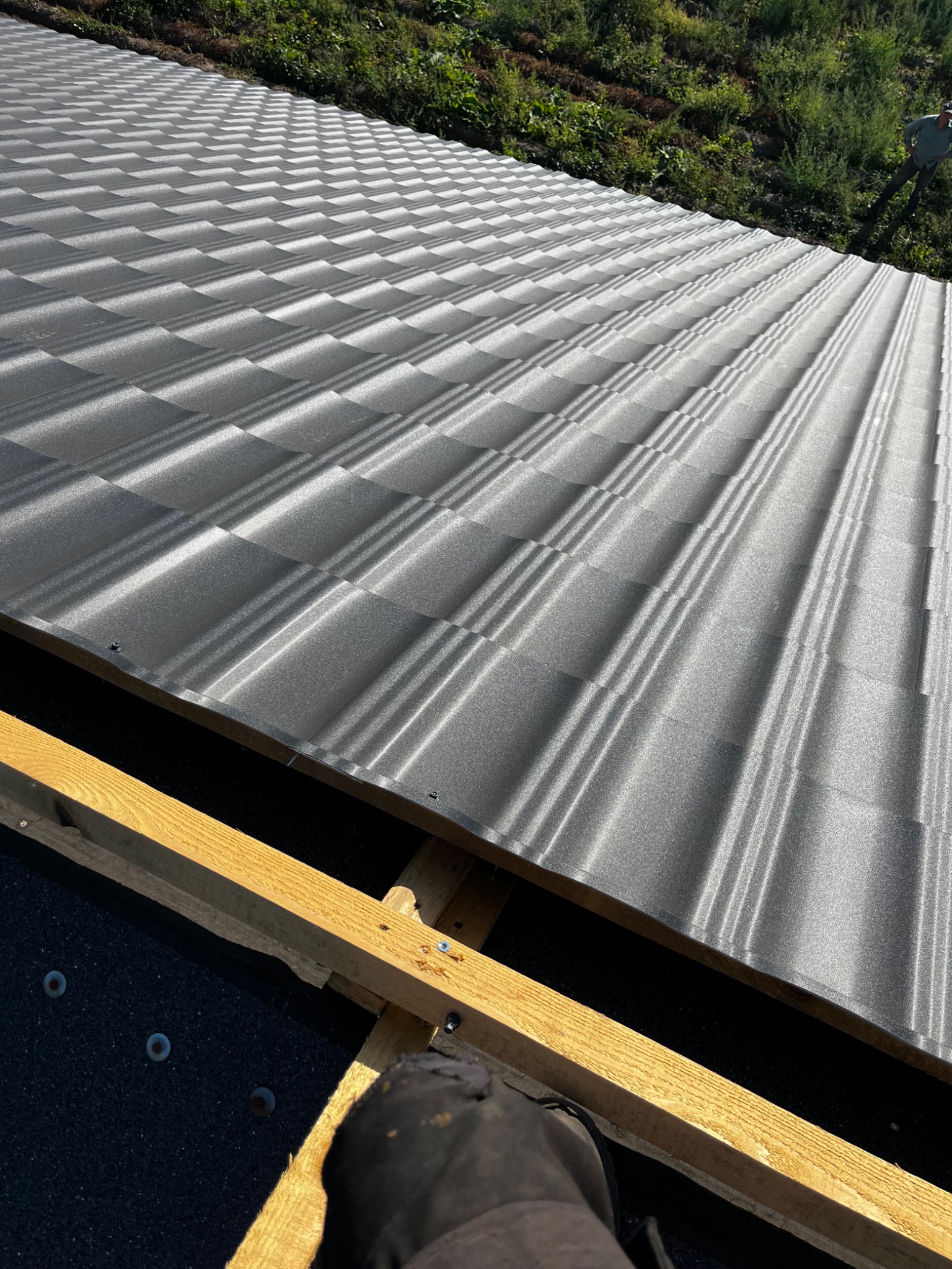 Szara blacha trapezowa na dachu w trakcie montażu, widoczne drewniane elementy konstrukcji i fragment buta dekarza w dolnym rogu. Ujęcie z góry.
