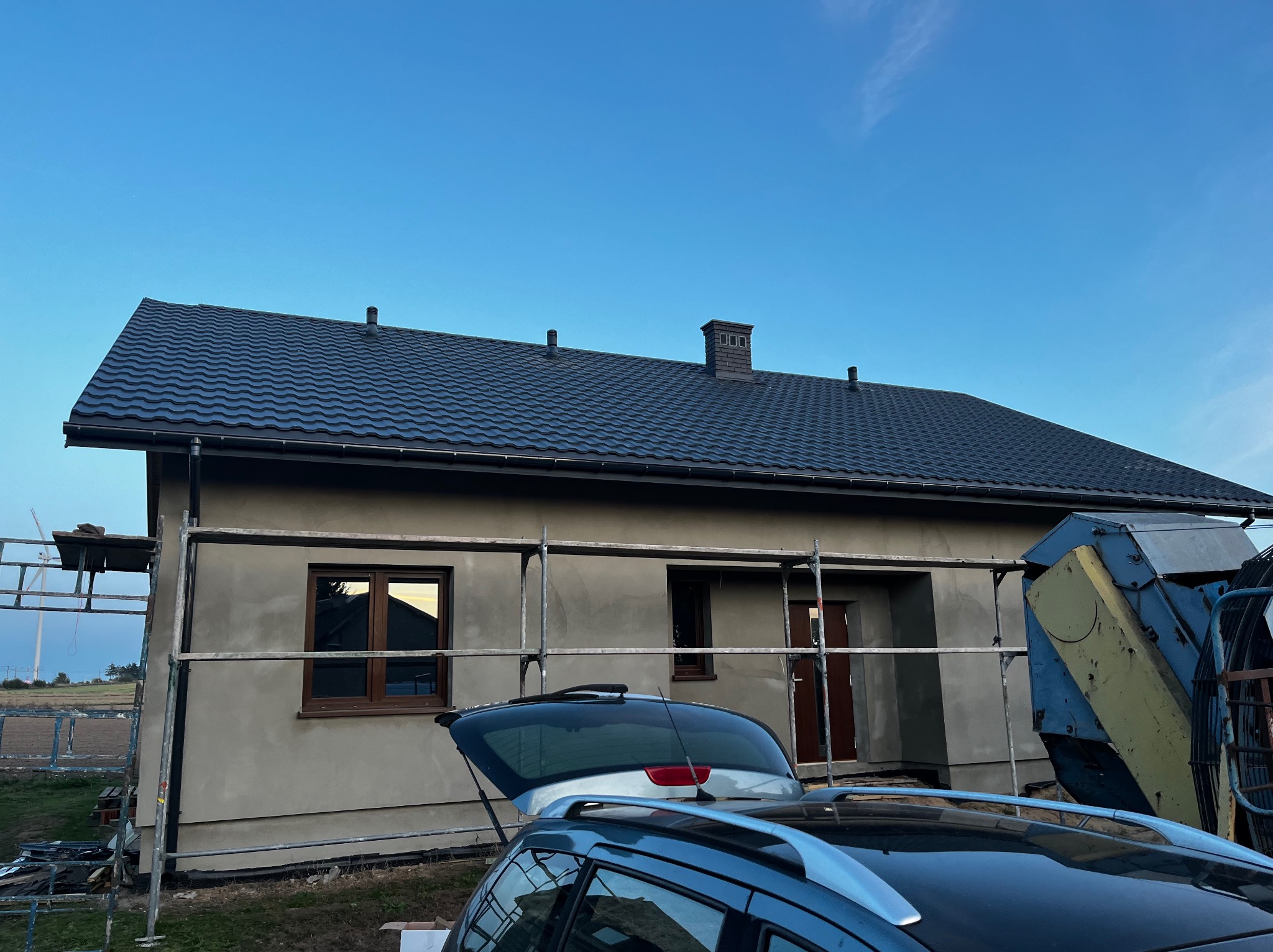 Dom jednorodzinny z ciemnym dachem z blachy, w trakcie prac wykończeniowych elewacji, z widocznym rusztowaniem i otwartym bagażnikiem samochodu na pierwszym planie.