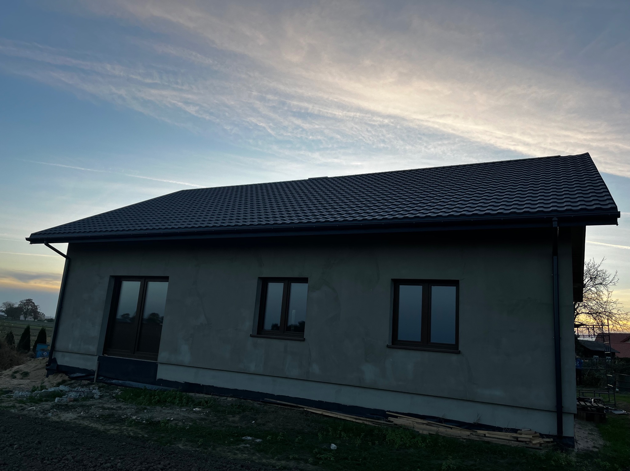 Nowoczesny dom jednorodzinny z ciemną, metalową dachówką w promieniach zachodzącego słońca. Widoczne rynny i surowe ściany zewnętrzne przed ociepleniem.
