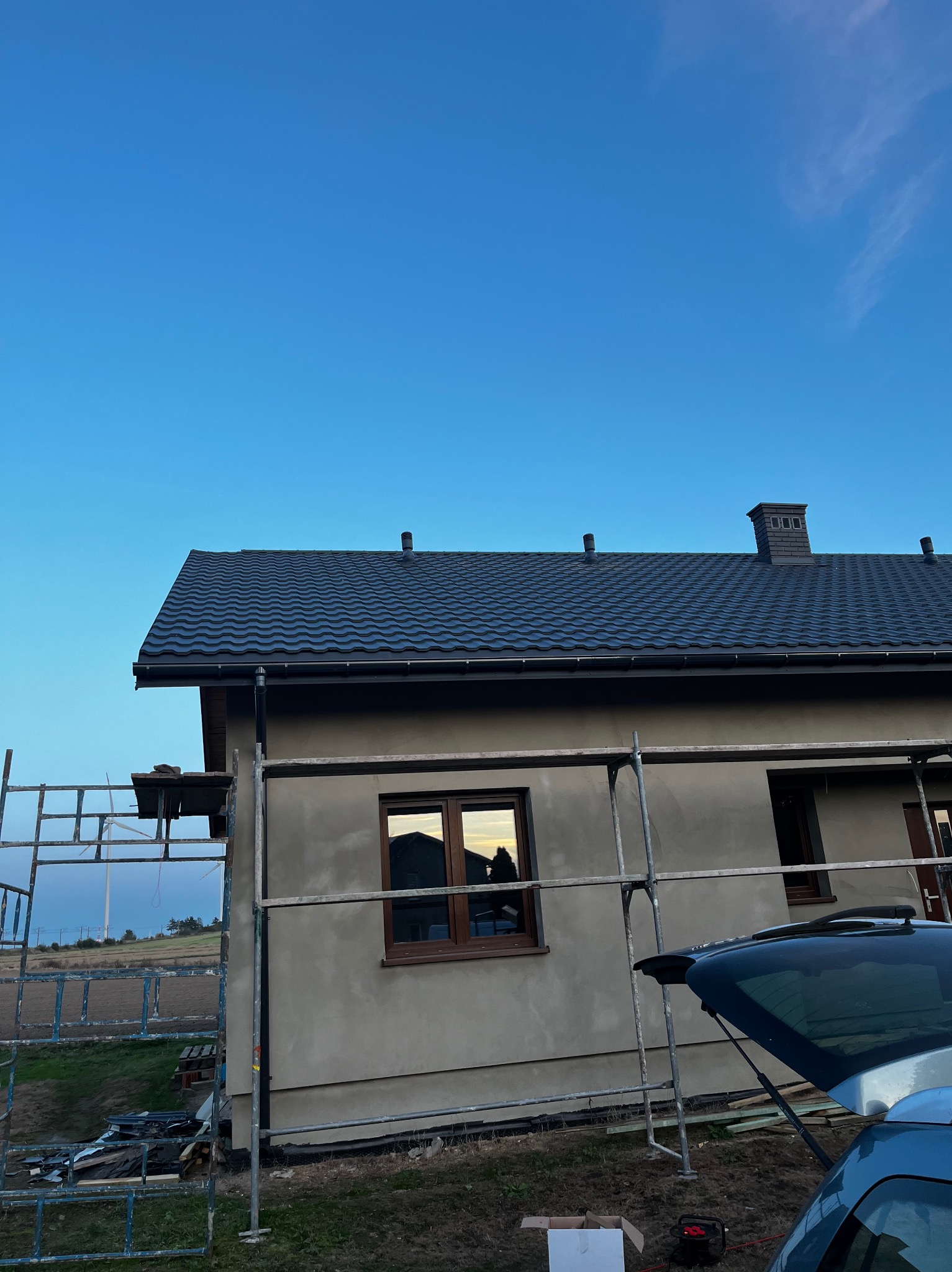 Dom z nowym, ciemnym dachem z blachodachówki, widoczny fragment rusztowania i otwarty bagażnik samochodu. W oknach odbija się zachodzące słońce.