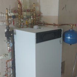 Usługi Hydrauliczne Matysik Patryk - Biały piec CO z miedzianą instalacją, zaworami i niebieskim zbiornikiem wyrównawczym na tle surowej ściany. Widoczne elementy instalacji grzewczej.