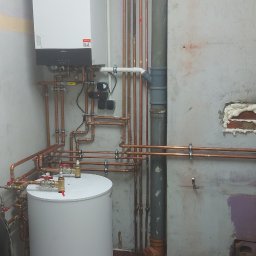 Usługi Hydrauliczne Matysik Patryk - Biały piec CO z podłączonymi miedzianymi rurami i zasobnikiem wody w pomieszczeniu z widocznymi instalacjami. Fragment ściany z uszczelnieniami pianką montażową.