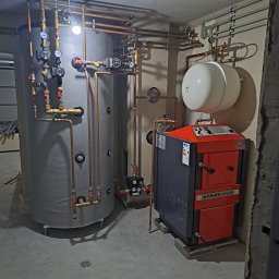 Usługi Hydrauliczne Matysik Patryk - Złożona instalacja grzewcza z kotłem Atmos i zasobnikiem, rury miedziane, manometry. Nowoczesny system w kotłowni, widok z dołu, szare ściany i podłoga.