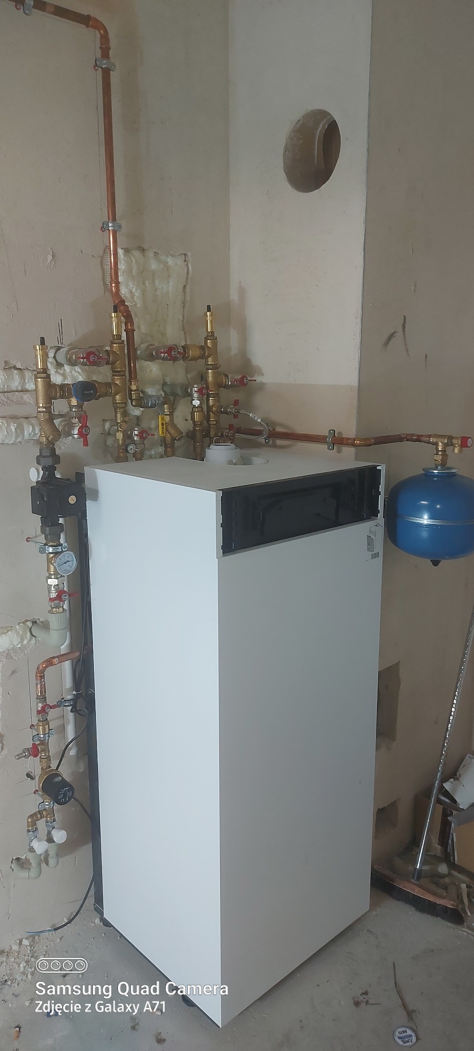 Biały piec CO z miedzianą instalacją, zaworami i niebieskim zbiornikiem wyrównawczym na tle surowej ściany. Widoczne elementy instalacji grzewczej.