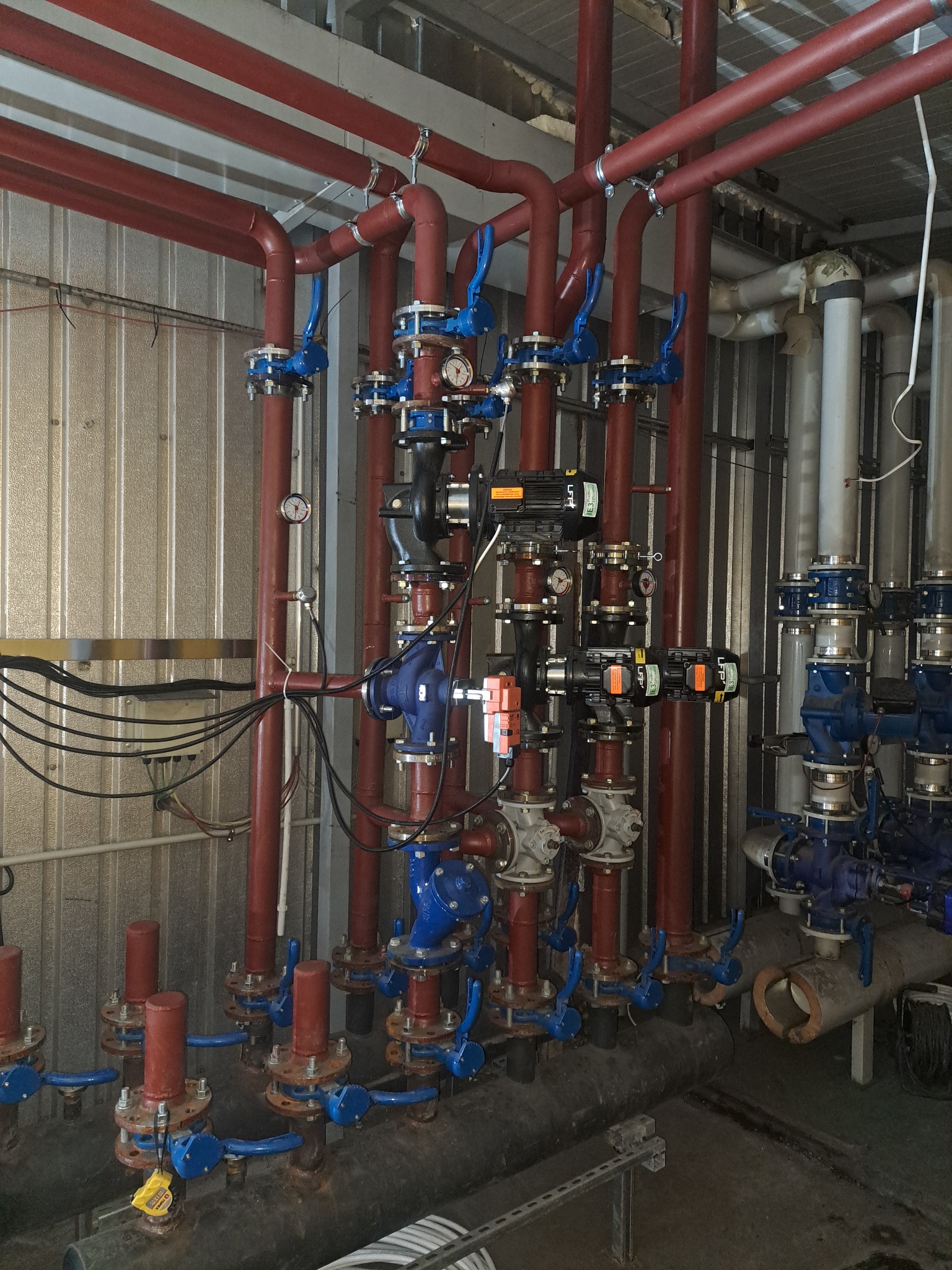 Rozbudowana instalacja rur w kolorach czerwonym i niebieskim, z zaworami i manometrami, na tle blaszanej ściany. Widoczne liczne połączenia i elementy armatury.