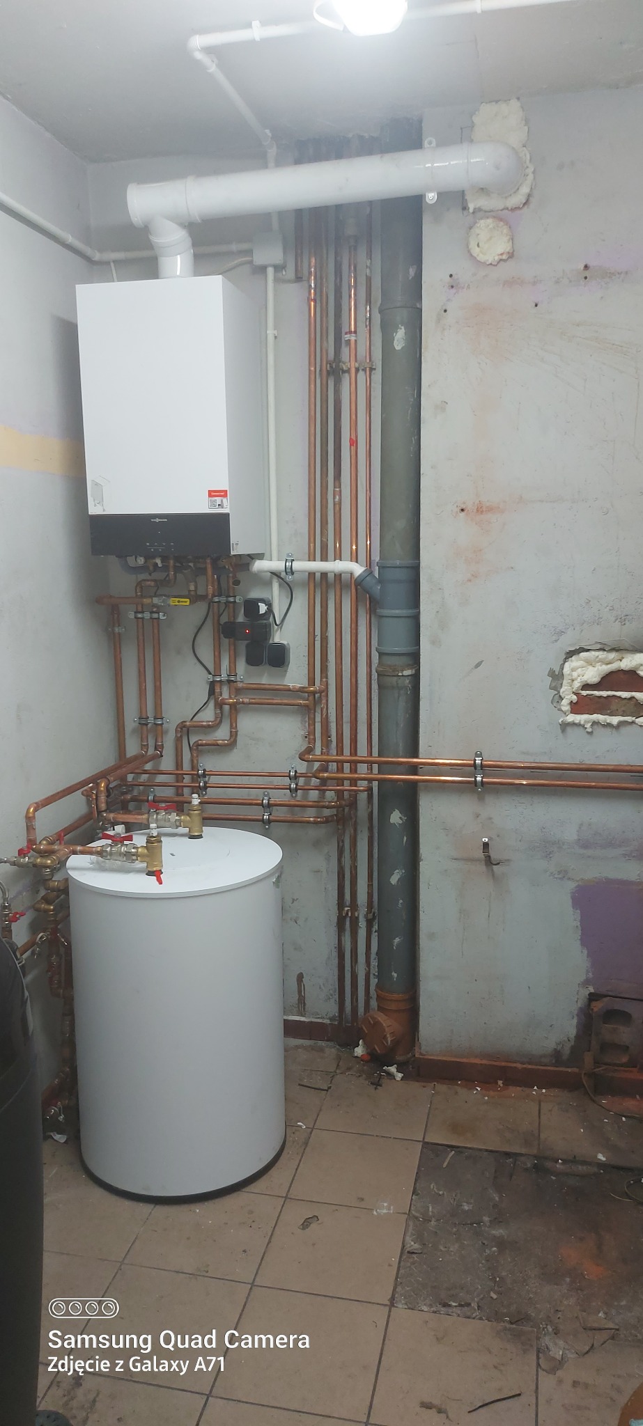 Biały piec CO z podłączonymi miedzianymi rurami i zasobnikiem wody w pomieszczeniu z widocznymi instalacjami. Fragment ściany z uszczelnieniami pianką montażową.