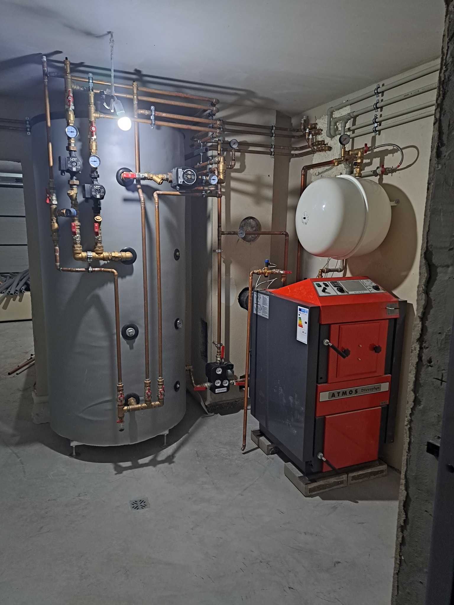 Złożona instalacja grzewcza z kotłem Atmos i zasobnikiem, rury miedziane, manometry. Nowoczesny system w kotłowni, widok z dołu, szare ściany i podłoga.