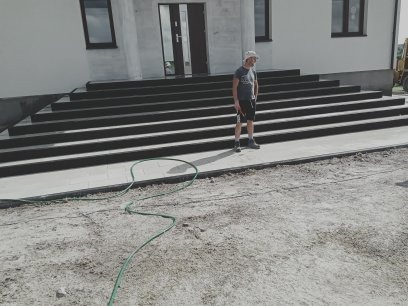 Nowoczesne schody wejściowe z ciemnego kamienia prowadzące do jasnego budynku, z ułożoną kostką brukową i osobą w tle. Wąż ogrodowy na pierwszym planie.