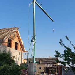 Complex dach - Wznoszenie więźby dachowej z użyciem dźwigu na tle błękitnego nieba. Widoczne elementy konstrukcyjne dachu i fragmenty muru. Prace dekarskie w toku.