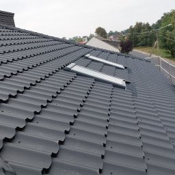 Complex dach - Czarny dach z blachodachówki z oknami dachowymi, montowany przez dekarza na rusztowaniu. Widok z góry, w tle zabudowa i zieleń.