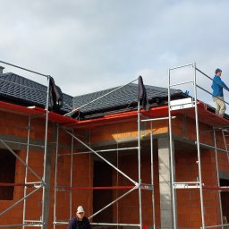 Complex dach - Dom w trakcie budowy z częściowo położonym, ciemnym dachem. Widoczne rusztowania i pracownicy. Dachówka ceramiczna, nowy system orynnowania w kolorze czerwonym.