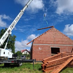 Complex dach - Biały dźwig Böcker podnosi impregnowaną drewnianą belkę na szczyt murowanego domu z charakterystycznym ceglanym szczytem, obok stos belek.