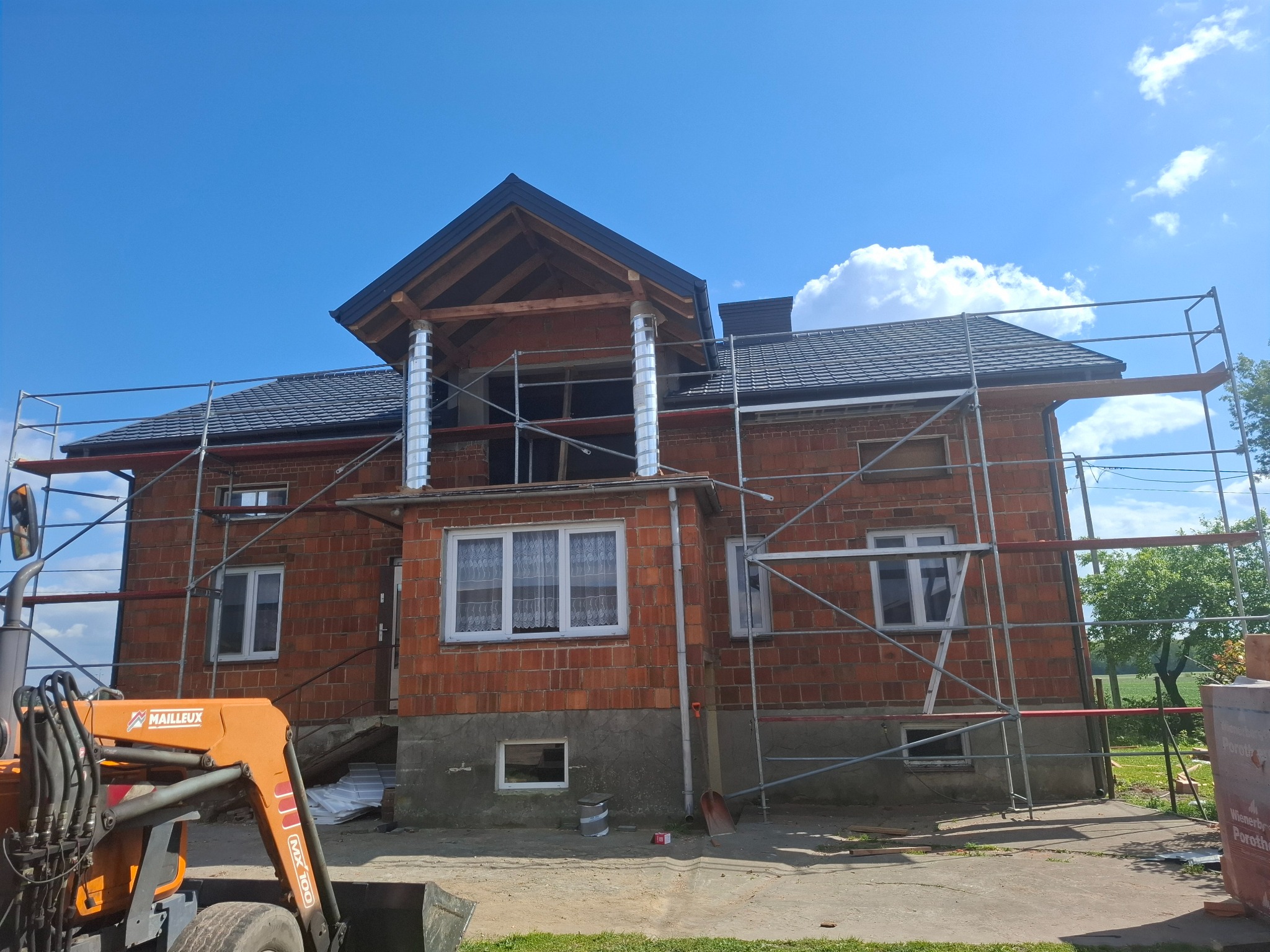 Dom w trakcie budowy w Dąbiu, z widocznym rusztowaniem, nowym dachem z ciemnej dachówki i kominami. Na pierwszym planie maszyna budowlana.