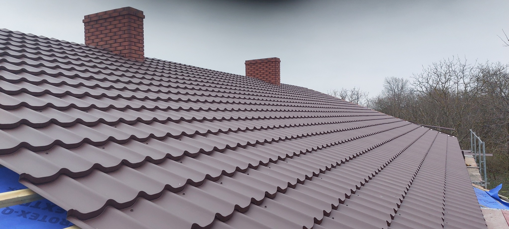 Brązowy dach z blachodachówki trapezowej, dwa kominy z cegły, widok z dołu pod kątem, drzewa w tle, niebieska membrana dachowa widoczna w dolnym rogu.