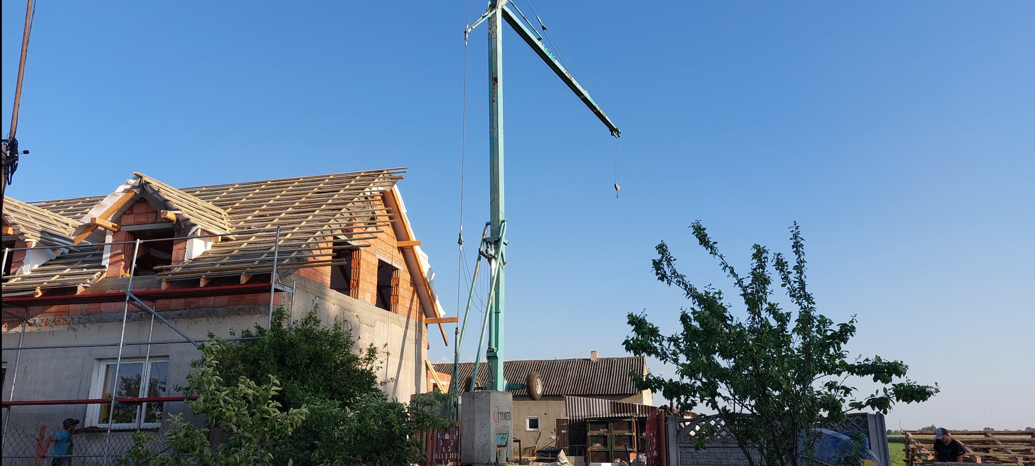 Wznoszenie więźby dachowej z użyciem dźwigu na tle błękitnego nieba. Widoczne elementy konstrukcyjne dachu i fragmenty muru. Prace dekarskie w toku.