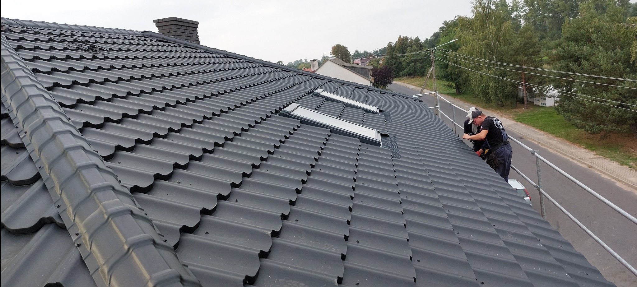 Czarny dach z blachodachówki z oknami dachowymi, montowany przez dekarza na rusztowaniu. Widok z góry, w tle zabudowa i zieleń.