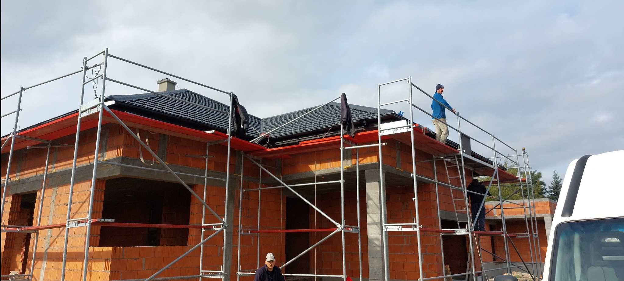 Dom w trakcie budowy z częściowo położonym, ciemnym dachem. Widoczne rusztowania i pracownicy. Dachówka ceramiczna, nowy system orynnowania w kolorze czerwonym.