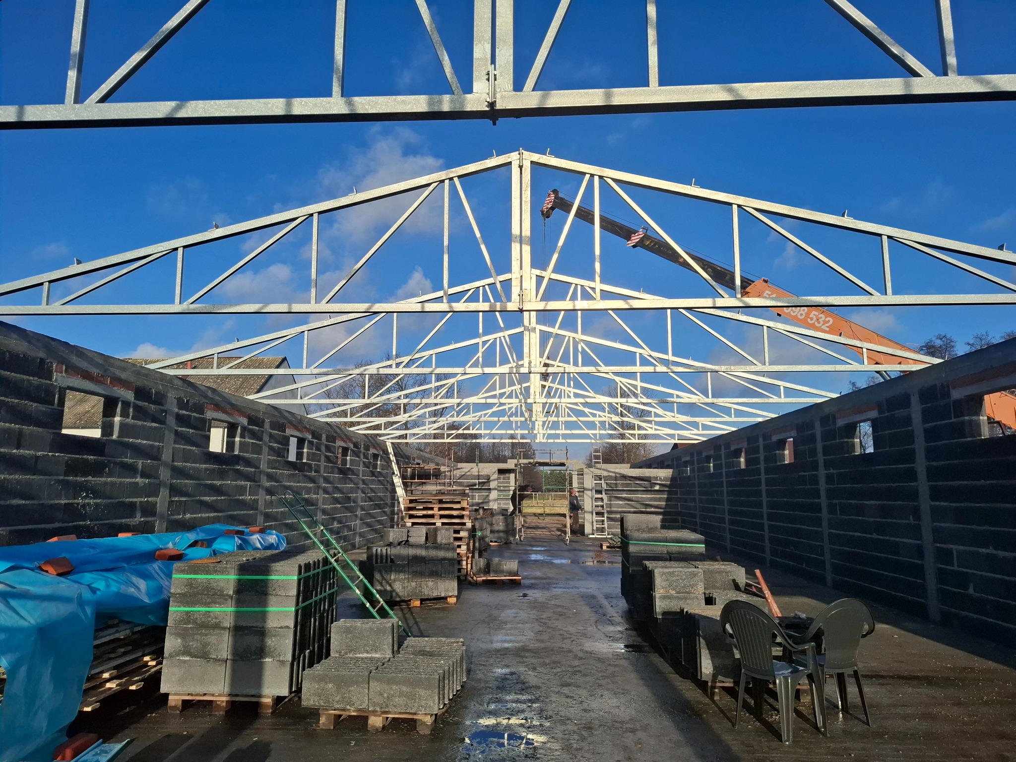 Wznoszenie konstrukcji dachu z metalowych elementów na budowie. Widoczny dźwig i materiały budowlane. Niebieskie niebo w tle.