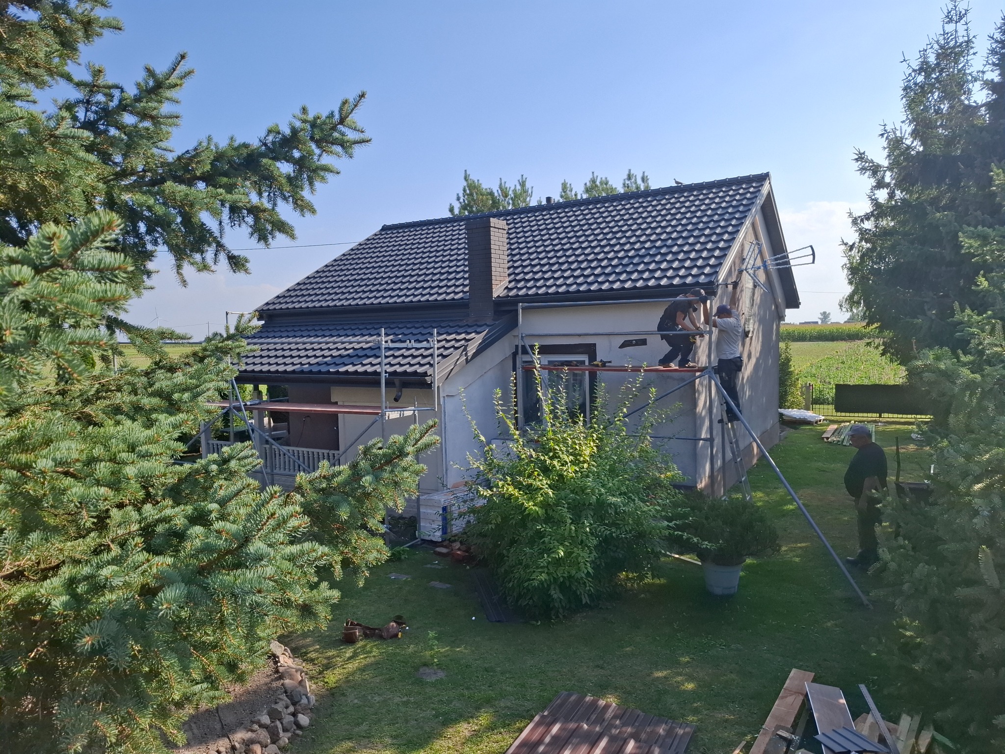 Dwóch monterów na rusztowaniu przy domu z ciemnym dachem, prawdopodobnie podczas prac dekarskich. Widok z góry, zieleń w tle.