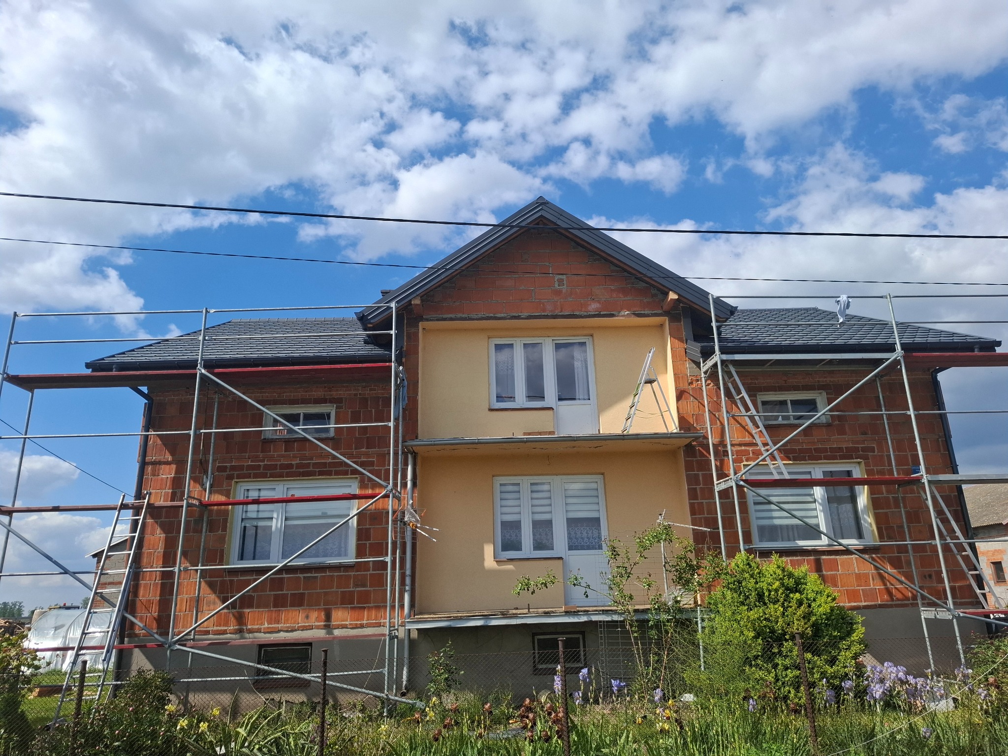 Dom w trakcie prac elewacyjnych z rusztowaniem. Część elewacji otynkowana na żółto, część z cegły. Dach pokryty szarą dachówką. Błękitne niebo z chmurami w tle.