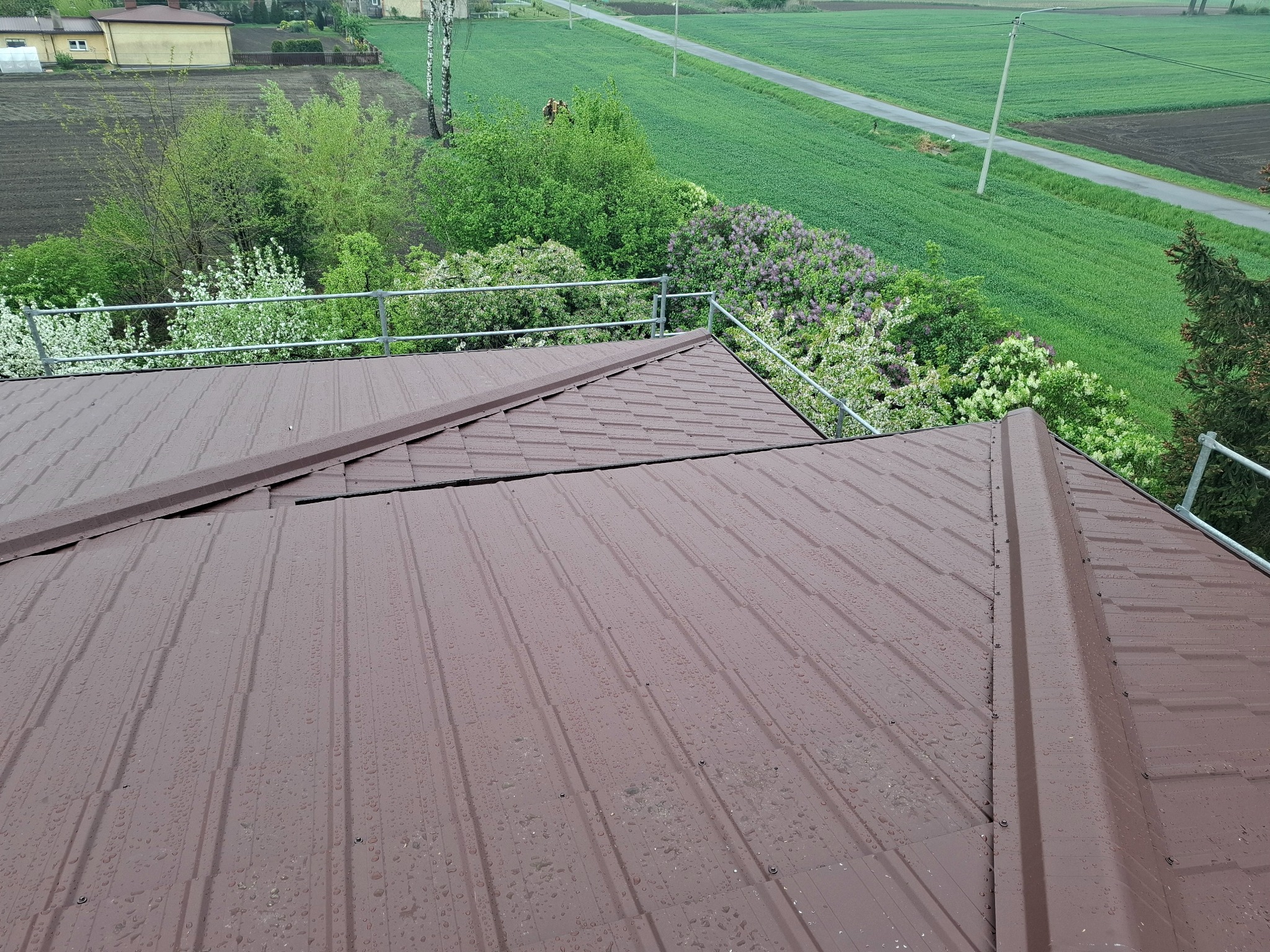 Ciemnobrązowe, metalowe pokrycie dachowe z widocznymi kroplami deszczu, z barierą ochronną i zielonym krajobrazem w tle. Widok z góry.