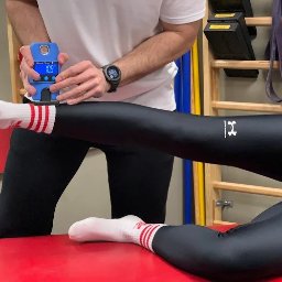 Fizjo-Fan - Fizjoterapia Masaż Trening medyczny - Pomiar siły mięśniowej uda pacjenta leżącego na czerwonym stole, za pomocą niebieskiego dynamometru. Widoczne drabinki gimnastyczne w tle.