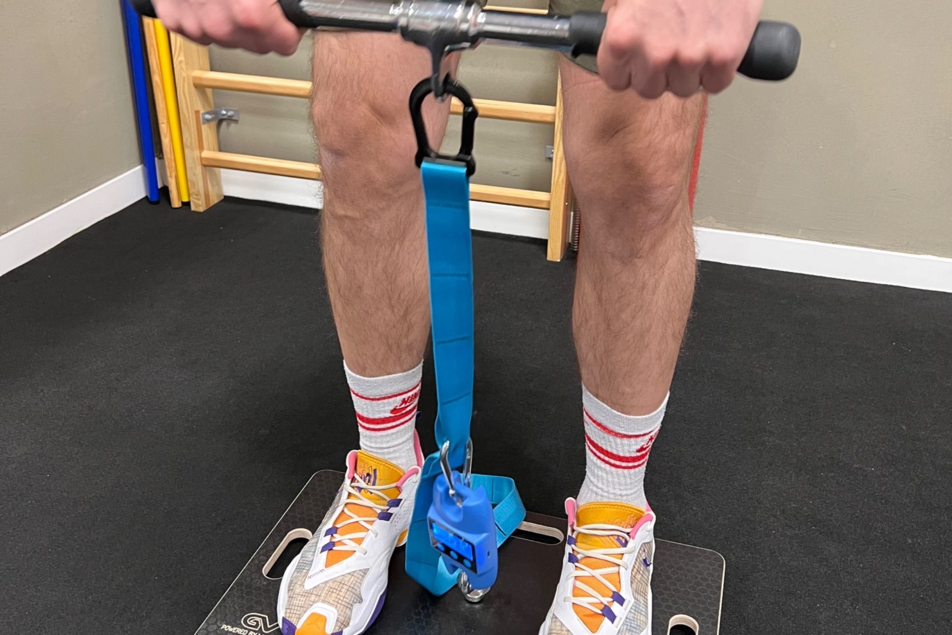 Osoba w sportowych butach i skarpetkach ćwiczy na platformie z użyciem specjalistycznego sprzętu do pomiaru siły mięśni nóg w gabinecie rehabilitacyjnym.