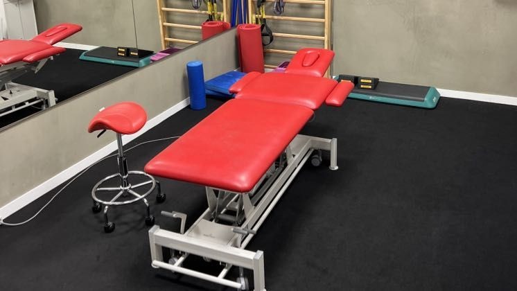 Czerwony stół do terapii manualnej z regulacją wysokości, obok krzesło siodłowe, drabinki gimnastyczne i stepy w jasnym gabinecie. Widok z boku.
