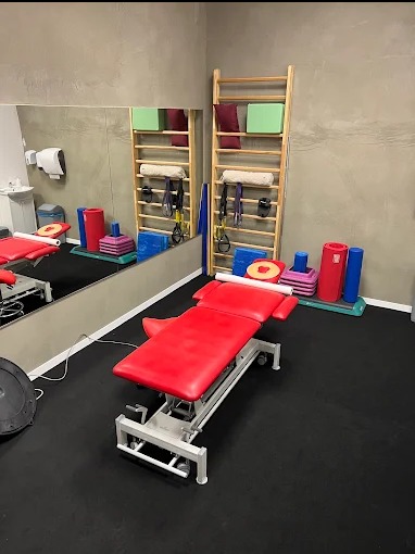 Jasne wnętrze gabinetu z czerwonym stołem do rehabilitacji na kółkach, drabinkami gimnastycznymi i lustrem. Widoczne akcesoria do ćwiczeń i czarna podłoga.