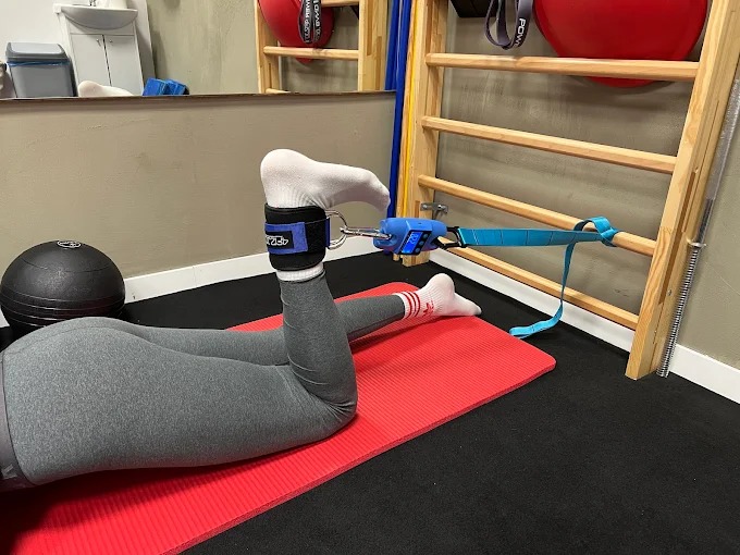 Kobieta w szarych legginsach ćwiczy na czerwonej macie z obciążeniem na kostce, przymocowanym do drabinki gimnastycznej. Widoczny ciężarek i piłka lekarska.