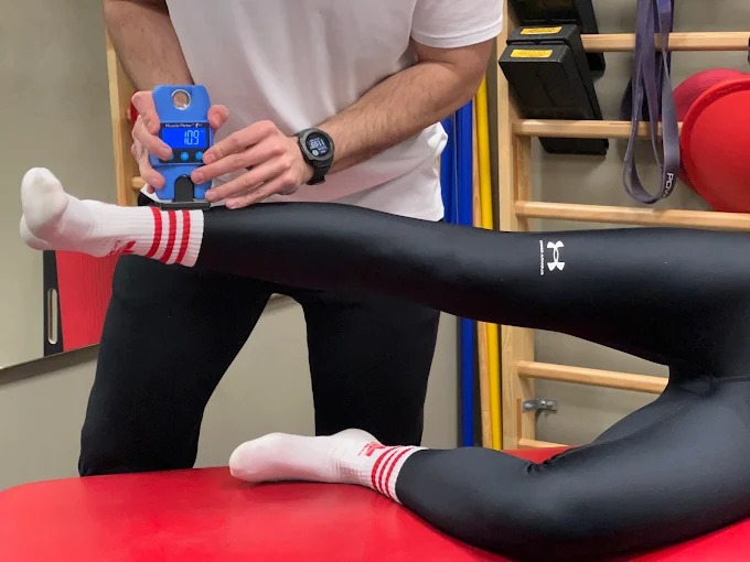 Pomiar siły mięśniowej uda pacjenta leżącego na czerwonym stole, za pomocą niebieskiego dynamometru. Widoczne drabinki gimnastyczne w tle.