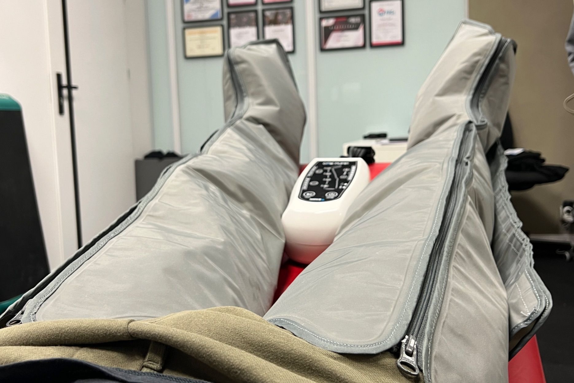 Nogi w mankietach do presoterapii z pilotem sterującym, widok z perspektywy pacjenta leżącego na czerwonym łóżku w gabinecie z certyfikatami na ścianie.