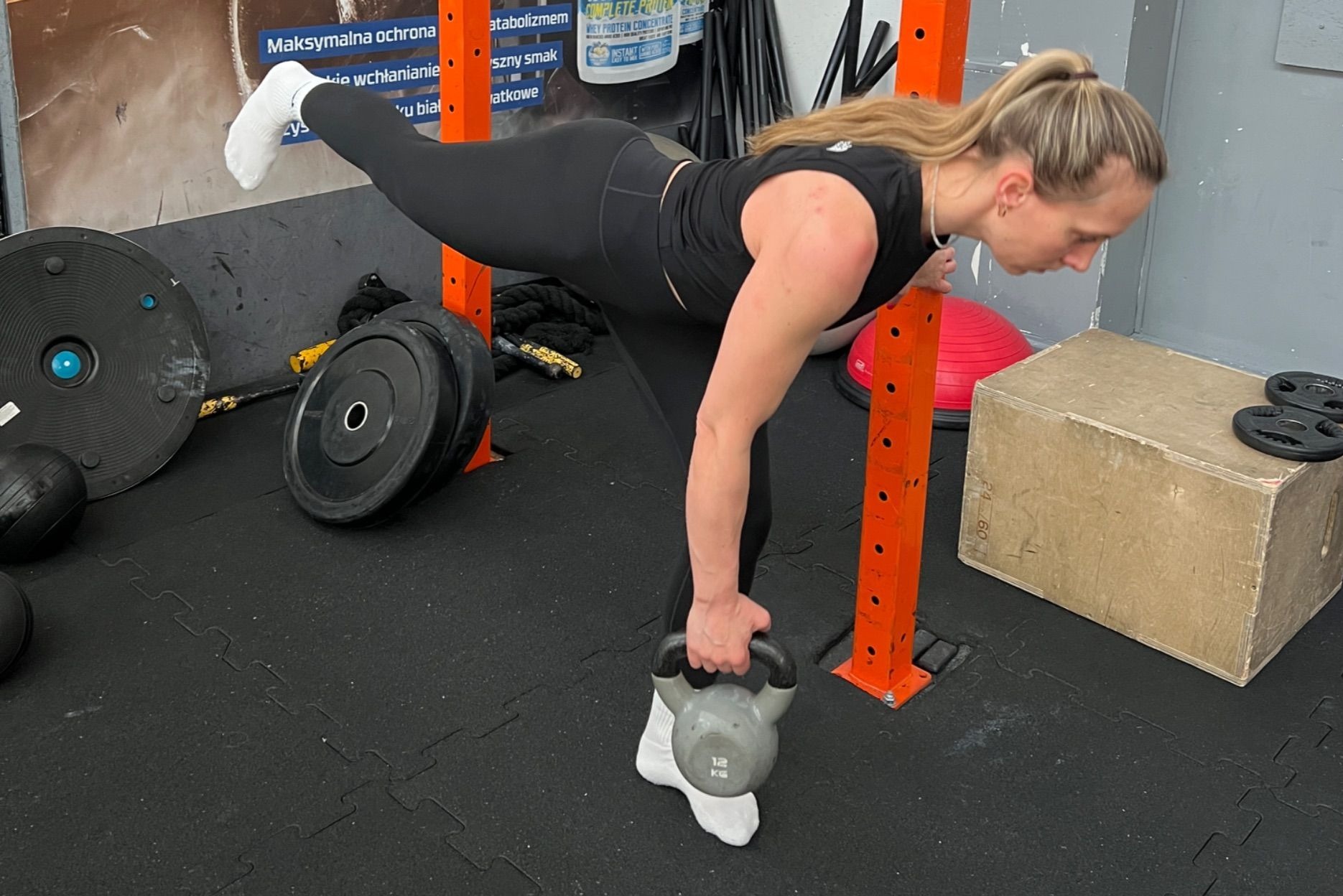 Kobieta w czarnym stroju ćwiczy równowagę z kettlebell 12kg w siłowni. Jedna noga uniesiona, druga podparta, ręka oparta o pomarańczową konstrukcję.