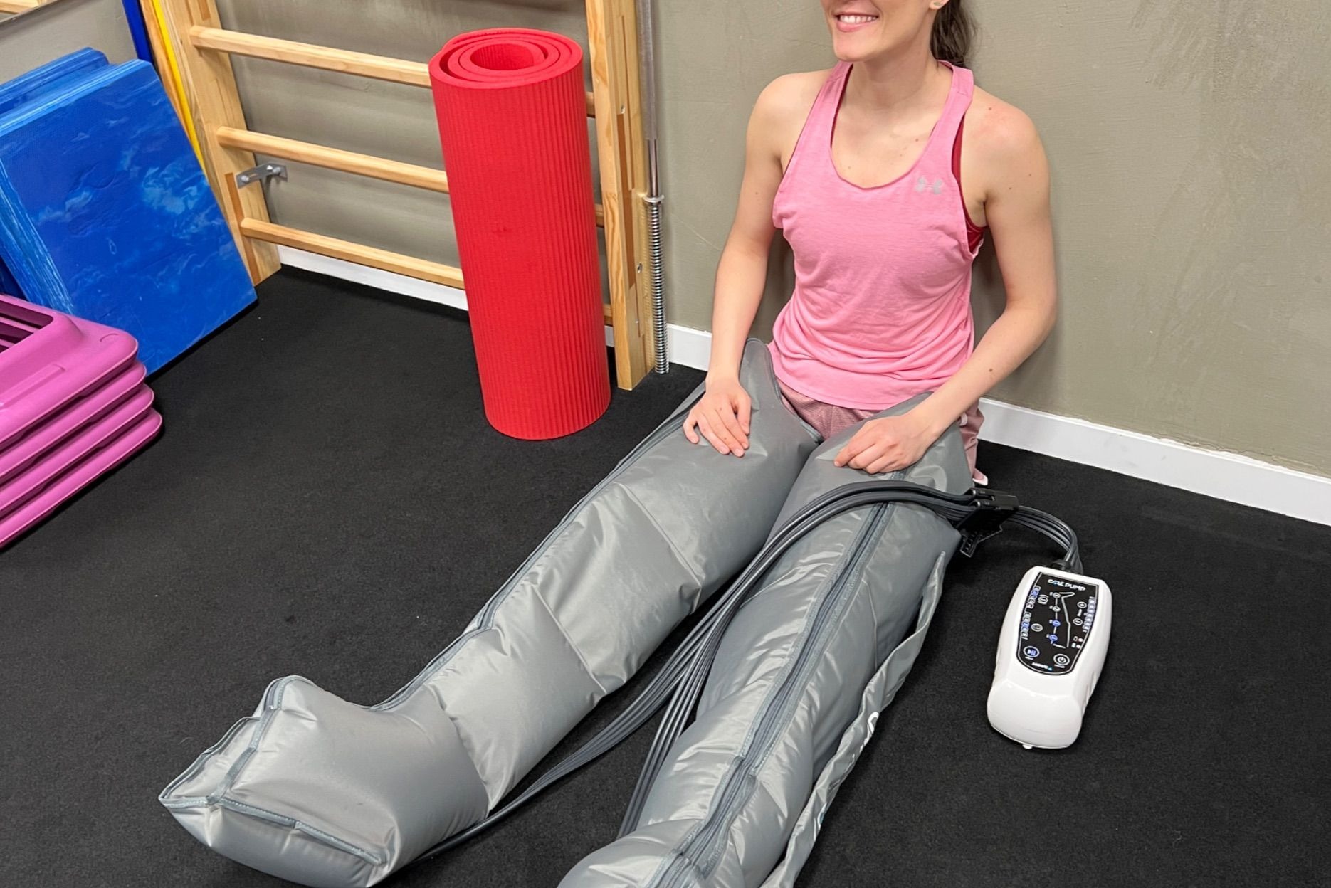 Kobieta siedzi z nogami w mankietach do presoterapii, obok leży urządzenie sterujące. W tle drabinki gimnastyczne i mata do ćwiczeń. Uśmiechnięta twarz.