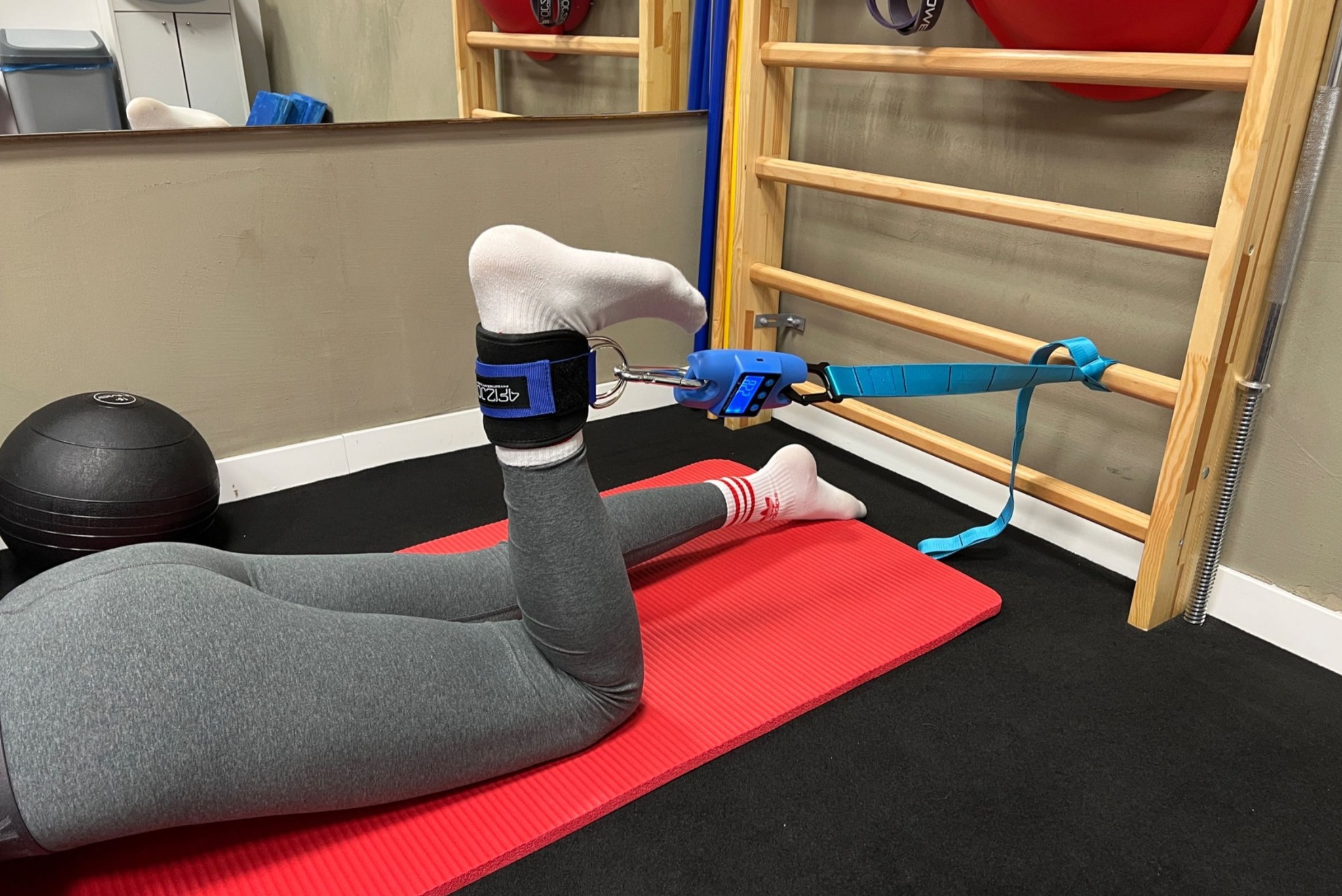 Osoba w szarych legginsach ćwiczy na czerwonej macie z użyciem specjalistycznego sprzętu do rehabilitacji nóg, przymocowanego do drabinki gimnastycznej.