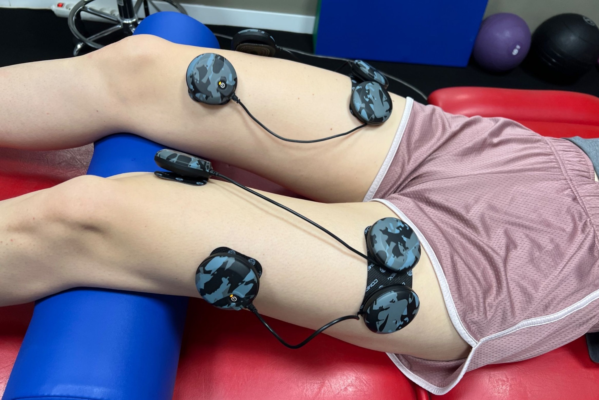 Osoba leży z elektrostymulacją na kolanach, na czerwonym podłożu. Widoczny niebieski wałek pod kolanami i piłki do ćwiczeń w tle. Elektrody w kamuflażu.