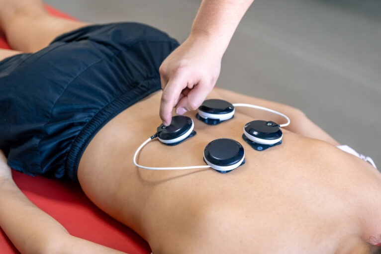 Pacjent leżący na brzuchu z elektrodami na plecach, dłoń terapeuty reguluje urządzenie. Zabieg elektrostymulacji w gabinecie rehabilitacji, widok z góry.