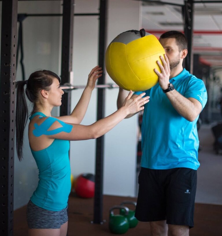 Kobieta z tapingiem na ramieniu i mężczyzna ćwiczą z żółtą piłką lekarską w siłowni. Trening funkcjonalny z obciążeniem w nowoczesnym fitness klubie.