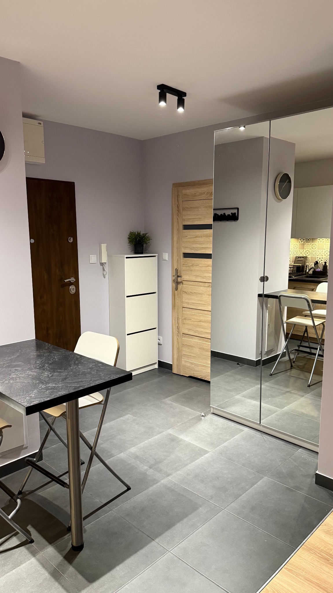 Nowoczesne wnętrze mieszkania z szarą podłogą z płytek, minimalistycznym stołem i lustrzaną szafą. Widoczne drzwi wejściowe i wewnętrzne, biała szafka na buty i oświetlenie sufitowe.