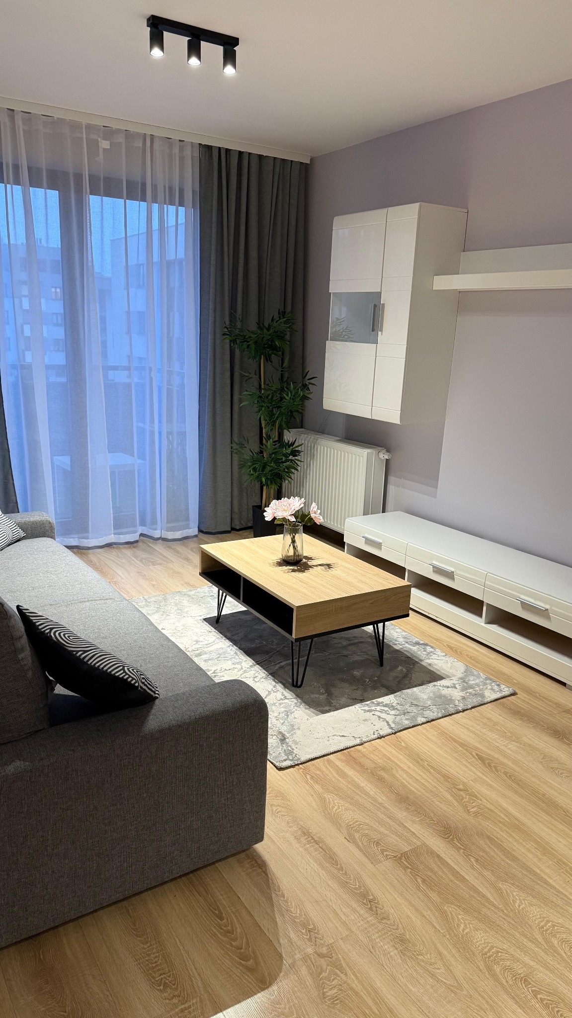 Jasne wnętrze salonu z szarą sofą, drewnianym stolikiem kawowym i białymi meblami na tle fioletowej ściany. Widoczne panele podłogowe i zasłony.