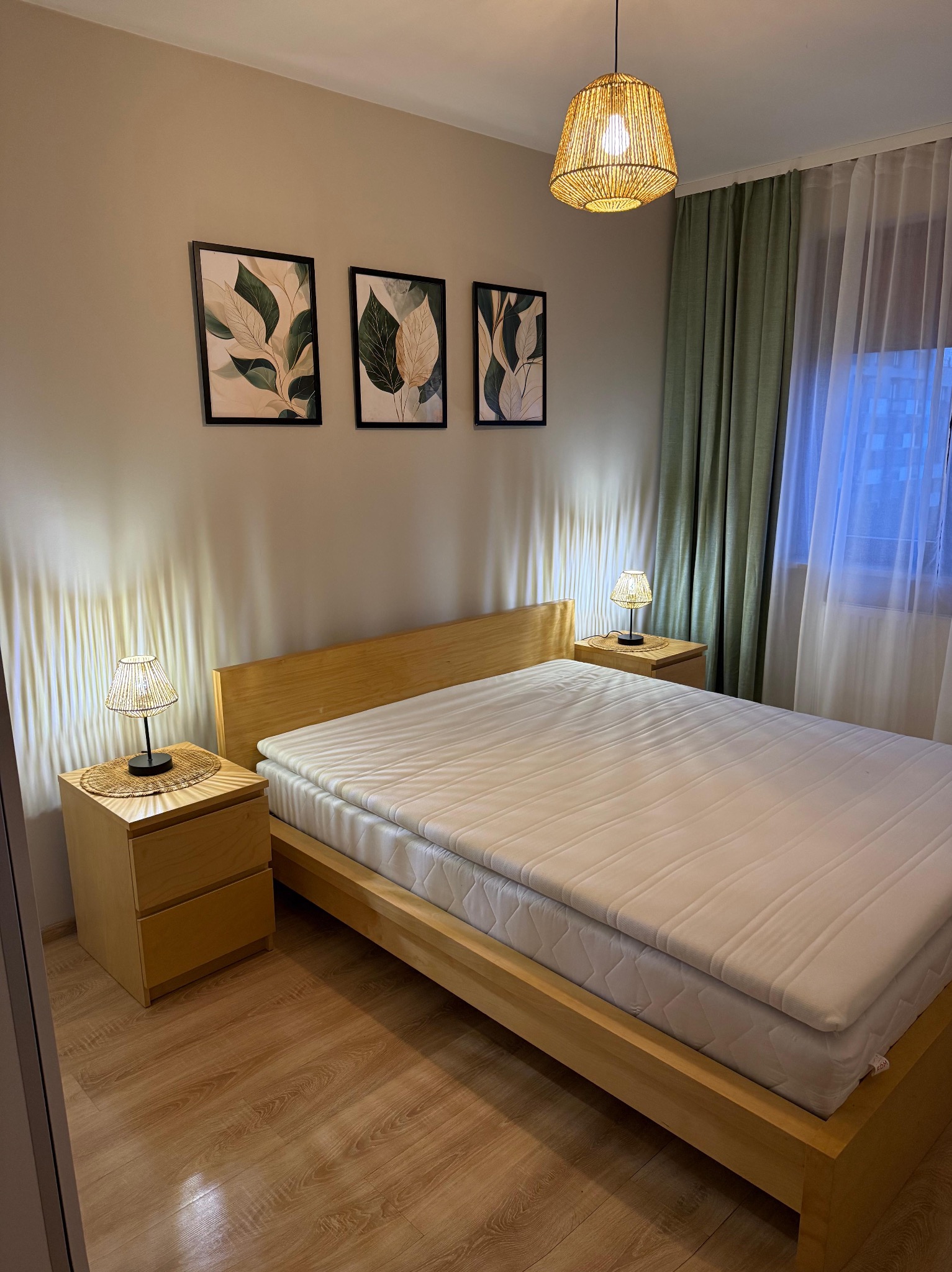 Jasna sypialnia z drewnianym łóżkiem, szafkami nocnymi i lampkami. Na ścianie trzy obrazy z motywem roślinnym. Okno zasłonięte firanką i zieloną zasłoną.