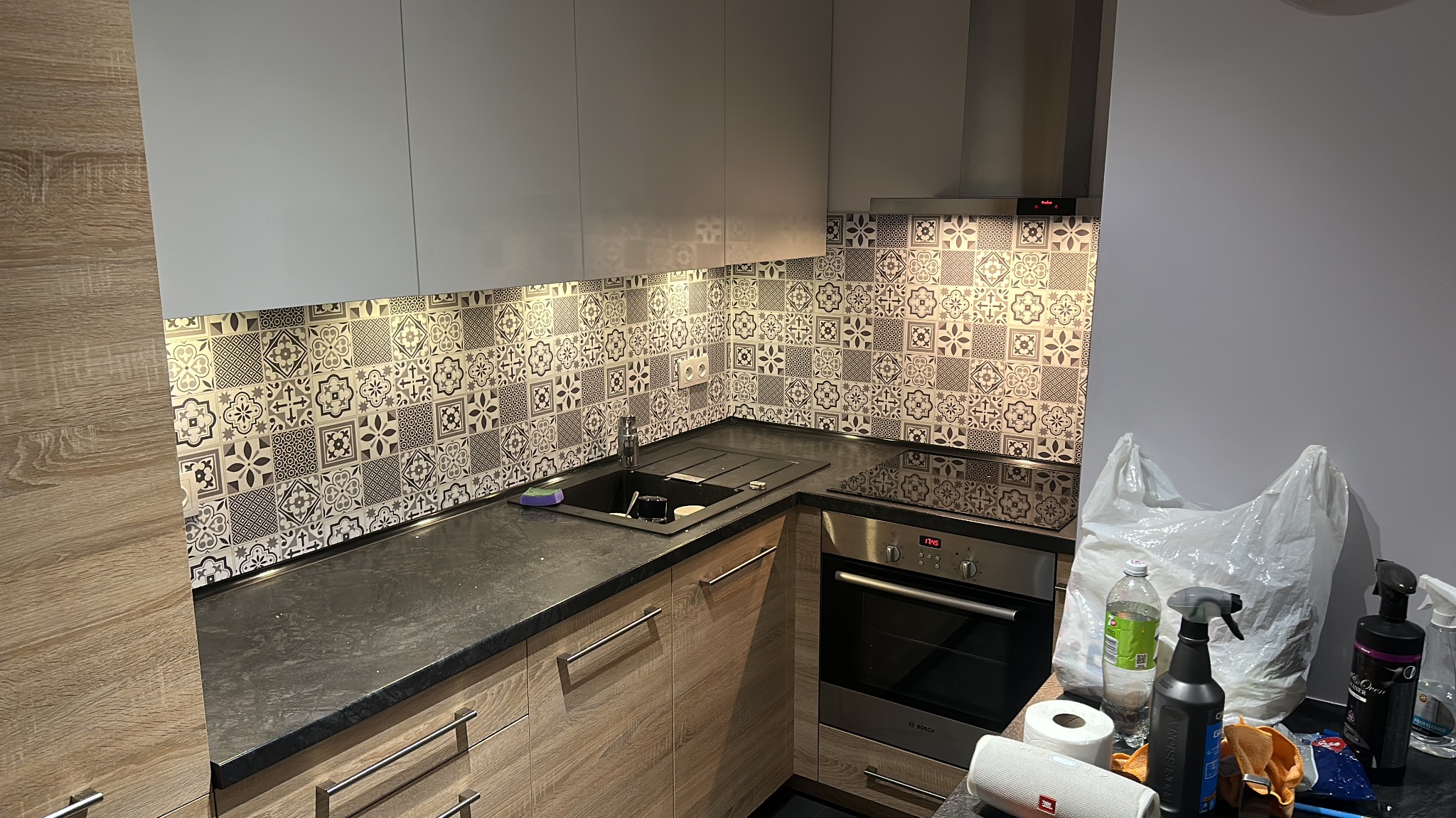 Kuchnia po remoncie: drewnopodobne szafki, czarny blat i dekoracyjne płytki na ścianie. Widoczne akcesoria kuchenne i środki czystości.