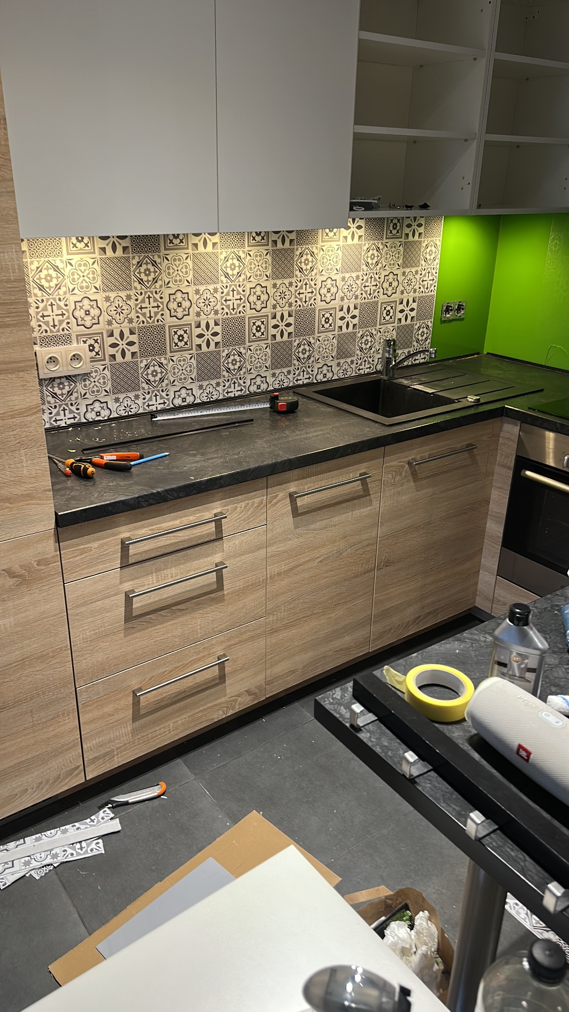 Kuchenna zabudowa z drewnopodobnymi frontami i czarnym blatem. Nad blatem dekoracyjne płytki. Widoczne narzędzia i akcesoria montażowe. Etap prac wykończeniowych.