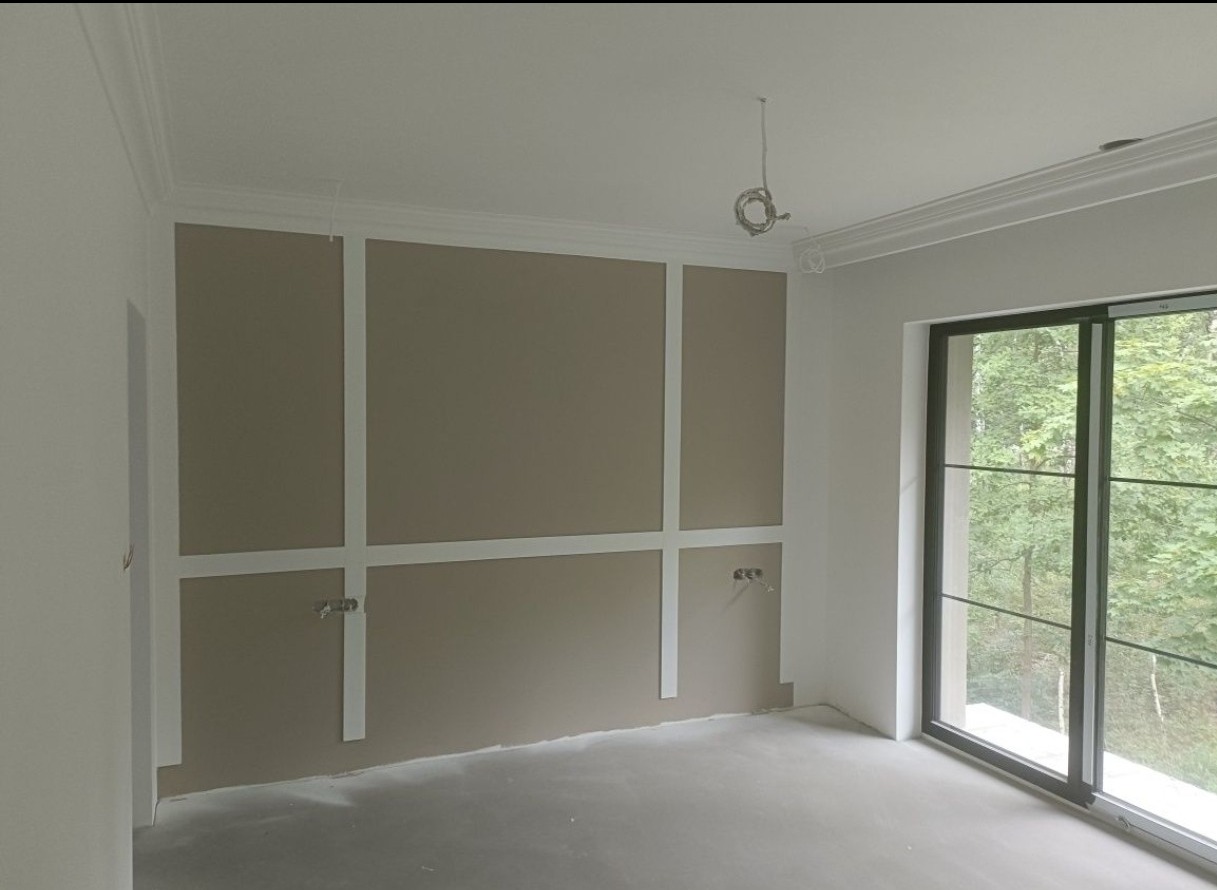 Pokój po szpachlowaniu z dekoracyjnymi listwami ściennymi i sztukaterią sufitową. Widoczne gniazdka elektryczne i duże okno przesuwne.