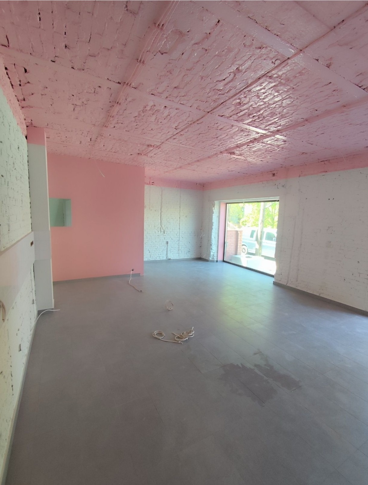 Surowe wnętrze po malowaniu: różowy sufit z cegły, białe ściany z widoczną fakturą, szara podłoga. Widoczne przewody elektryczne. Duże okno w tle.